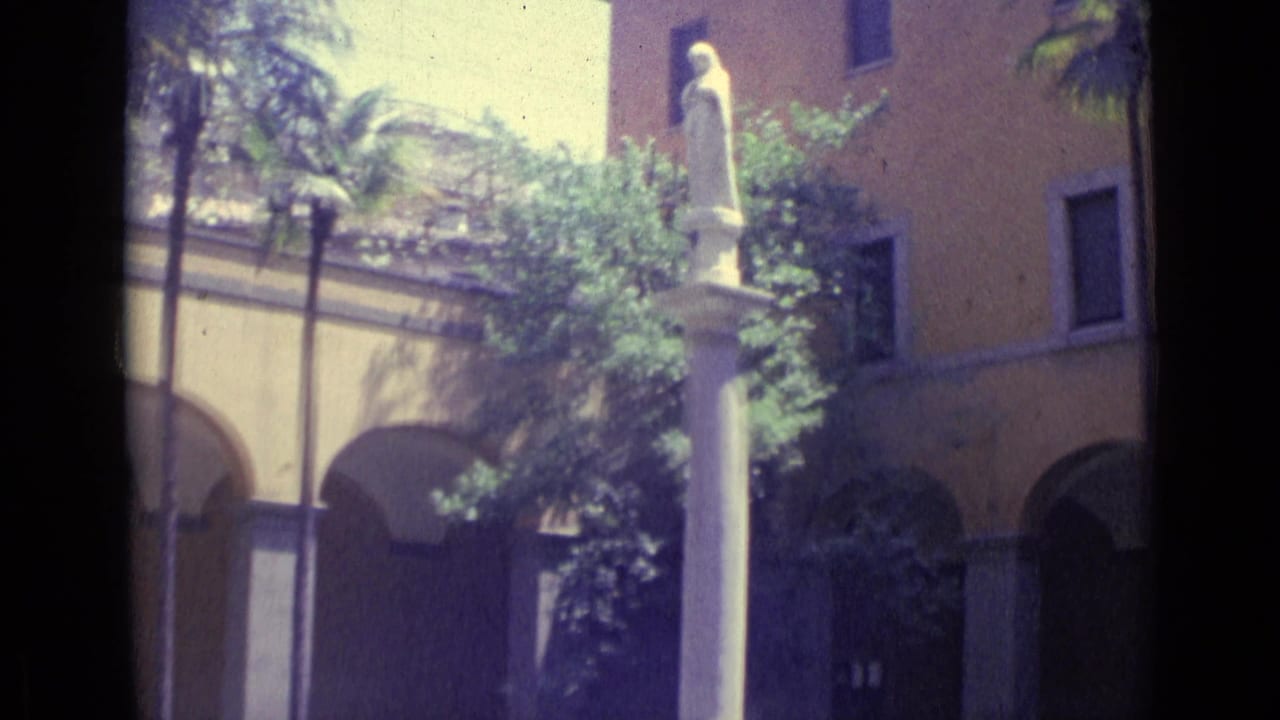 Small white statue on a tall column in a sunlit courtyard with palm trees and arched yellow walls.