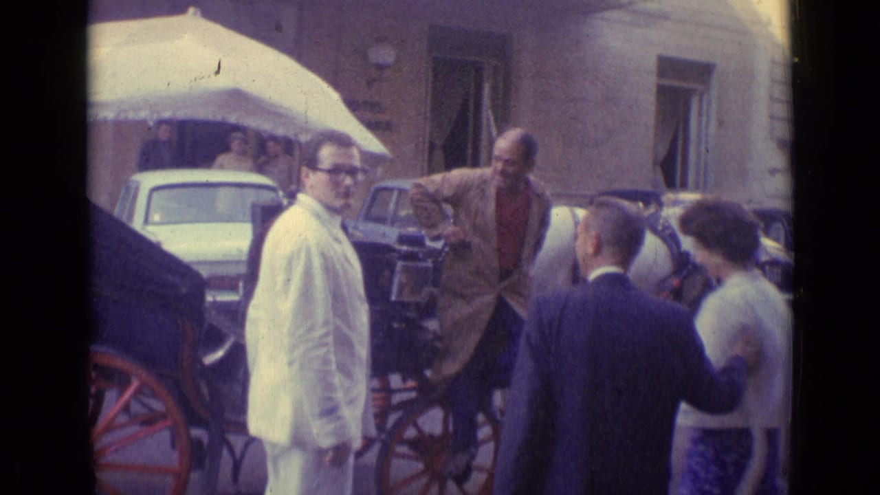 1962 Vatican City frame showing suited men and a woman near a canopied carriage, with parked cars and courtyard windows behind.