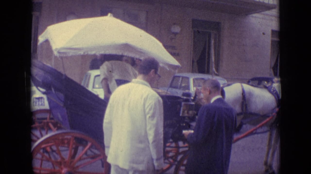 A man in a white suit and a man in a dark suit stand near a carriage horse and umbrella-covered seat.