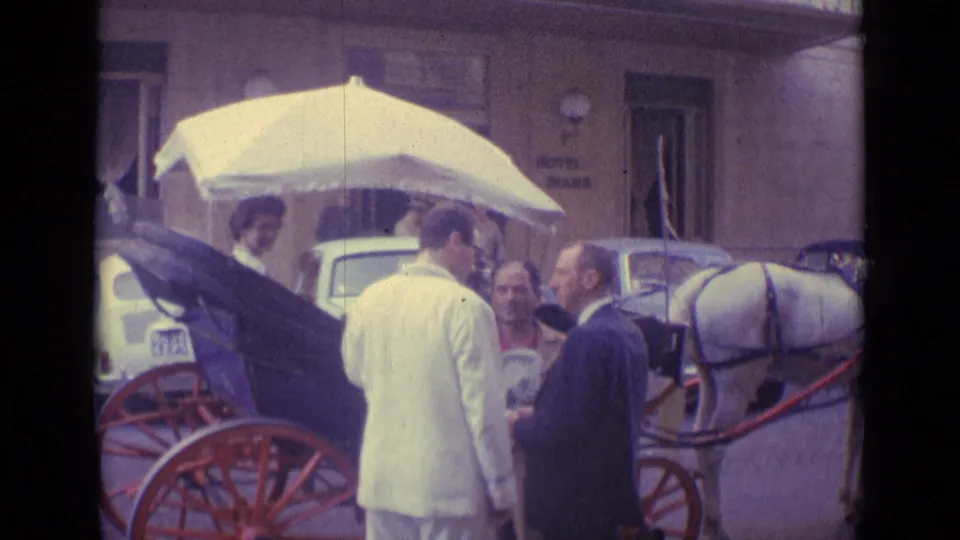 1962 Vatican City street frame with adults beside a horse-drawn carriage, a seated woman, red wheels, and hotel signage.