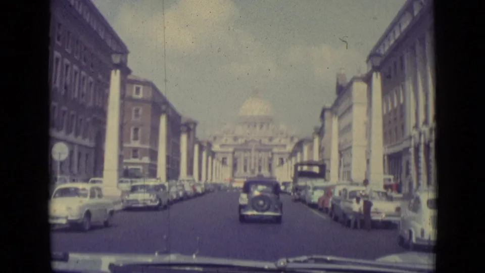 Moving-vehicle view down a Vatican City street toward St. Peter's Basilica, with parked classic cars and traffic ahead.