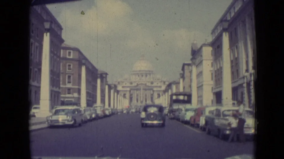 1962 moving-car view along a Vatican City boulevard with period cars, pedestrians, and St. Peter's Basilica ahead.