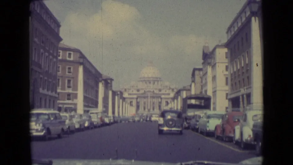 Forward view from a car on a Vatican City street, with parked 1960s automobiles, pedestrians, and St. Peter's Basilica in the distance.