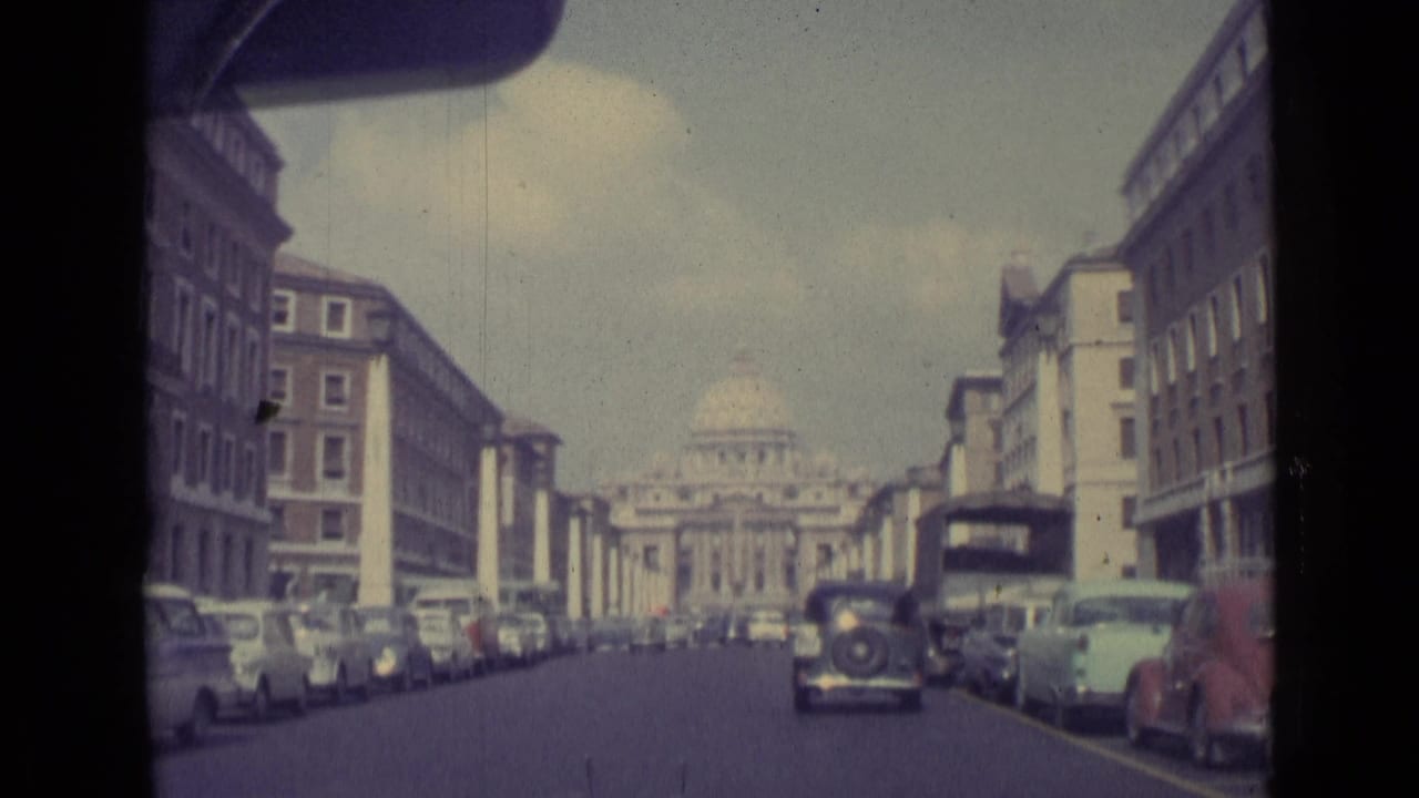 1962 Vatican City home-movie frame showing a car approach along a broad avenue toward St. Peter's Basilica, with parked cars on both sides.