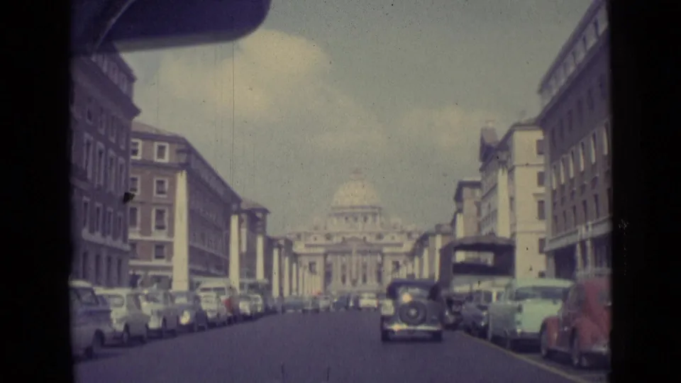 1962 Vatican City home-movie frame showing a car approach along a broad avenue toward St. Peter's Basilica, with parked cars on both sides.