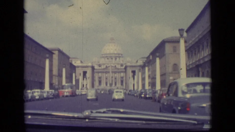 1962 Vatican City frame from inside a car, showing the dashboard foreground, avenue traffic, columns, and St. Peter's Basilica ahead.