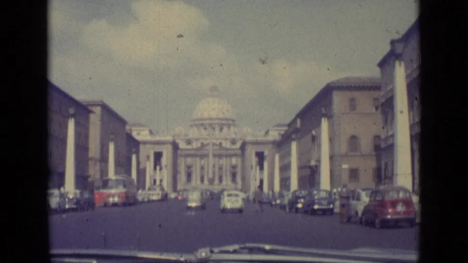 1962 home-movie view down a Vatican City avenue, with early 1960s cars and St. Peter's Basilica centered beyond the street.
