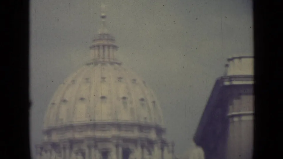 1962 Vatican City frame with St. Peter's Basilica dome centered and a nearby building edge at right, softened by 8mm film grain.