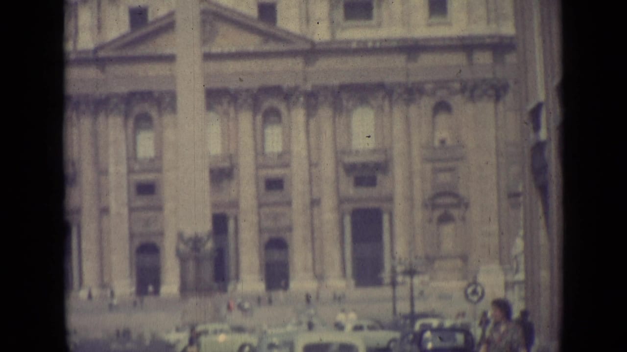 1962 Vatican City home-movie frame showing a grand stone facade with columns and small figures near vehicles in the square.