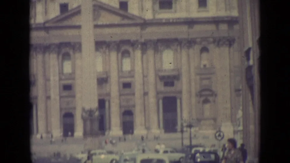 1962 Vatican City home-movie frame showing a grand stone facade with columns and small figures near vehicles in the square.