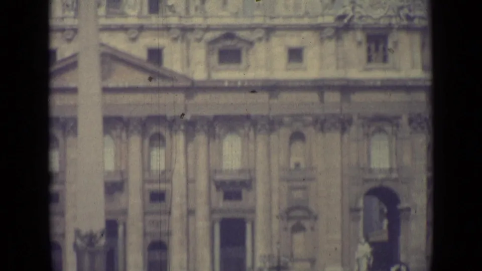1962 Vatican City home-movie frame showing the lower facade, a statue near an arched entrance, and cars along the square.