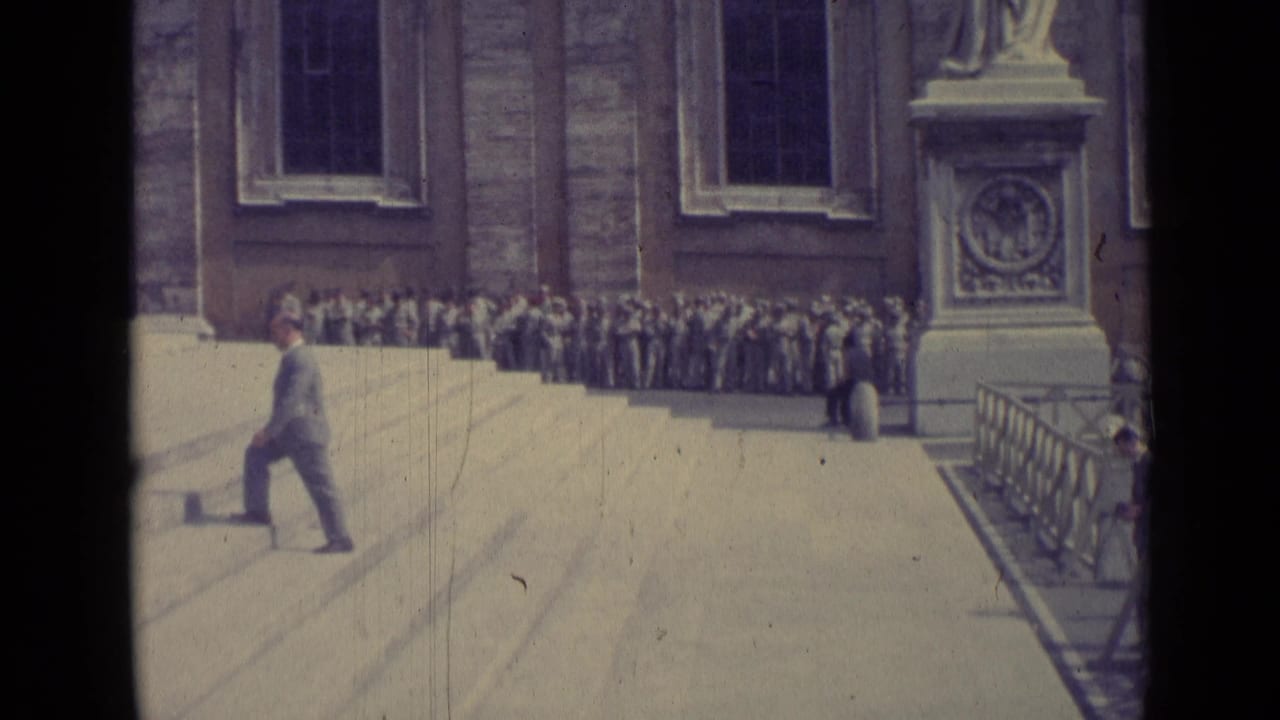 Man in a suit walks across broad steps near a crowd gathered by a large stone building.