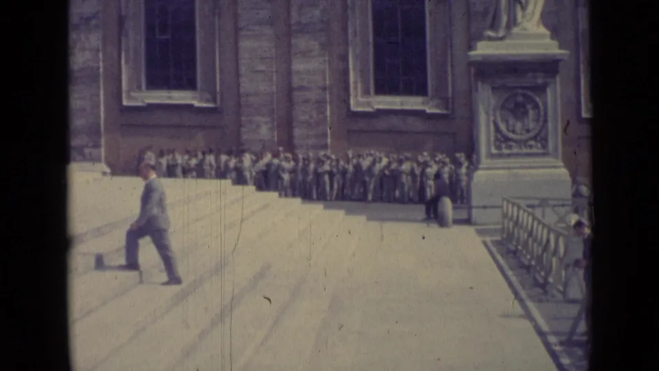 1962 Vatican City frame with a suited man crossing the steps as a crowd stands near tall windows and a statue pedestal.