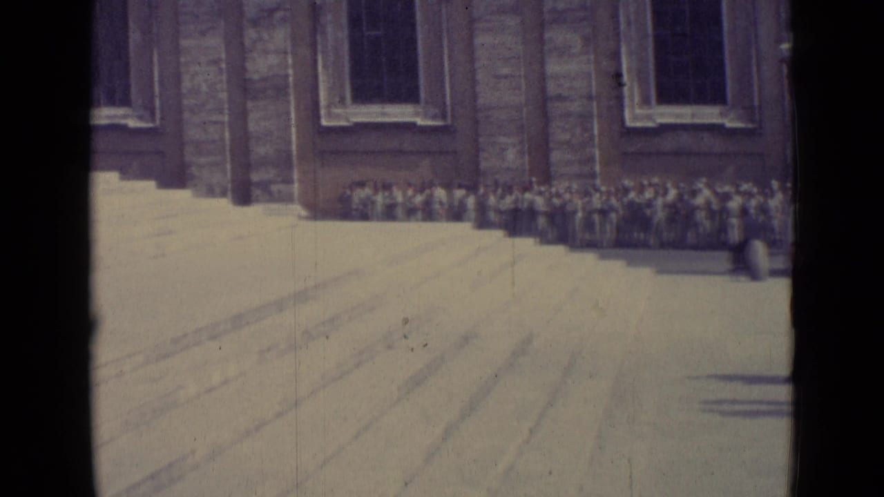 1962 Vatican City frame with a wide view of people gathered along the building wall below tall windows and broad open steps.