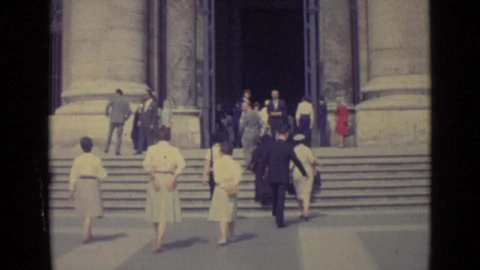 1962 Vatican City frame showing visitors in formal dress moving toward a basilica entrance with broad steps and columns.
