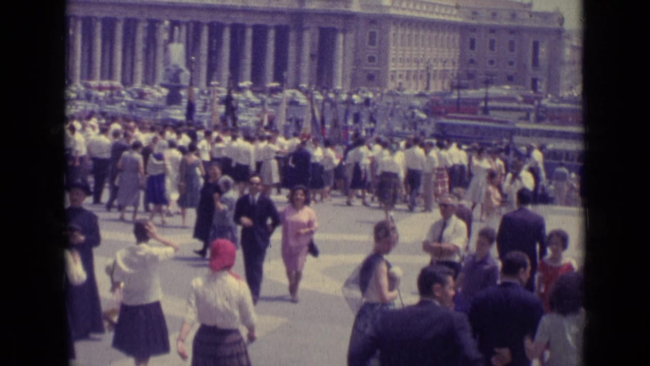 Crowd crossing a broad plaza in front of a large colonnaded building.