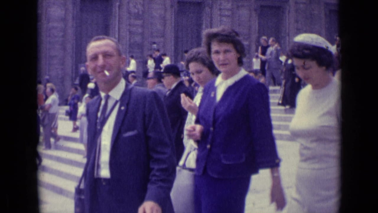 1962 Vatican City frame with a smoking man in a dark suit and women in blue and white clothing near broad stone steps.