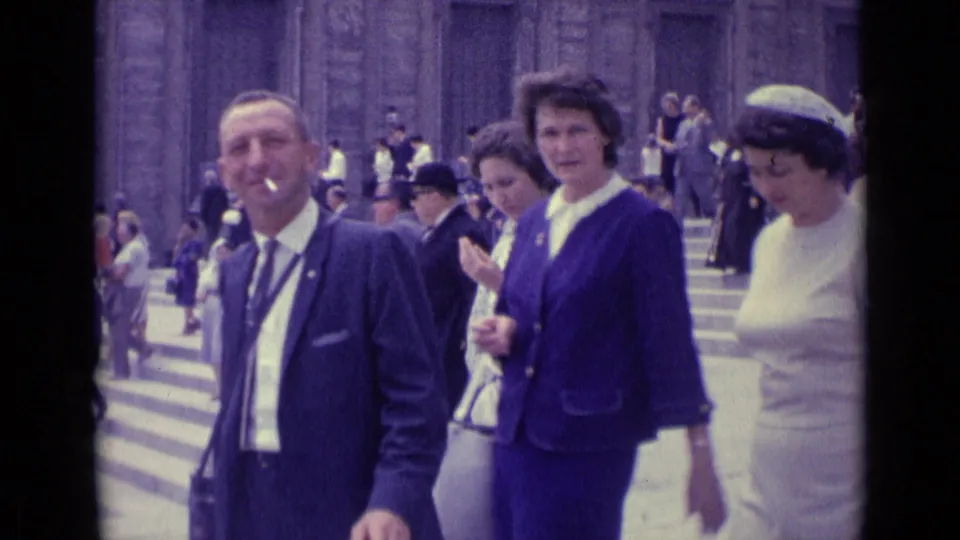 1962 Vatican City frame with a smoking man in a dark suit and women in blue and white clothing near broad stone steps.