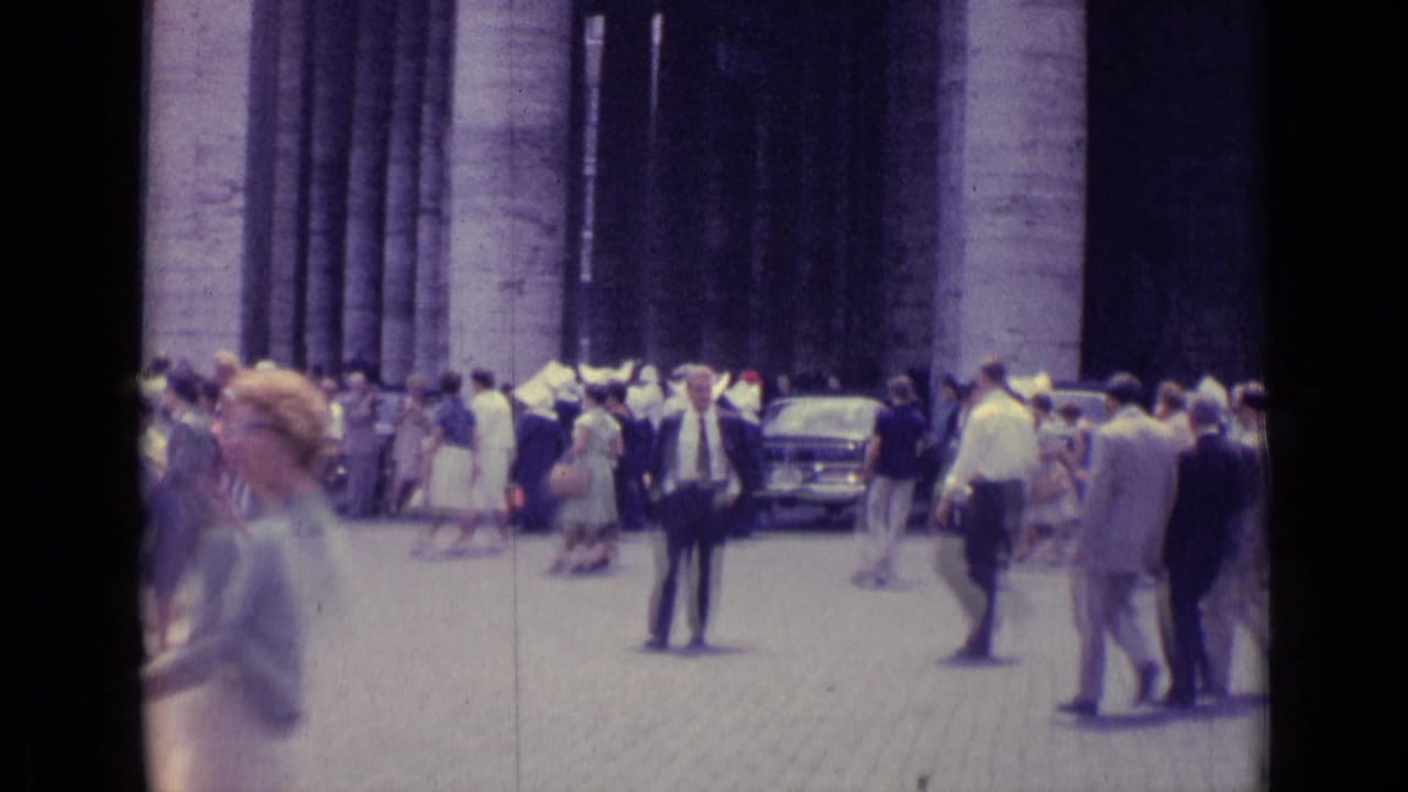Crowd of people walking near tall stone columns with parked cars in the background.