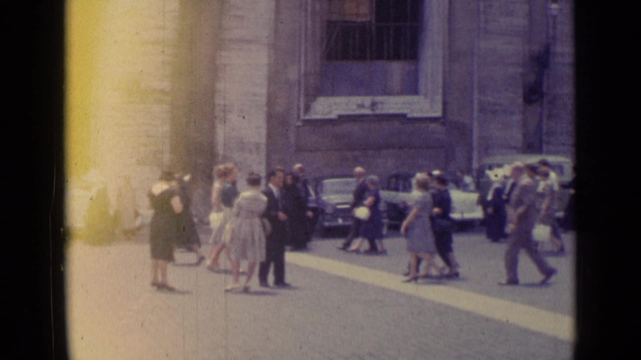 1962 Vatican City frame showing people crossing a bright paved square near parked cars and a stone building facade.