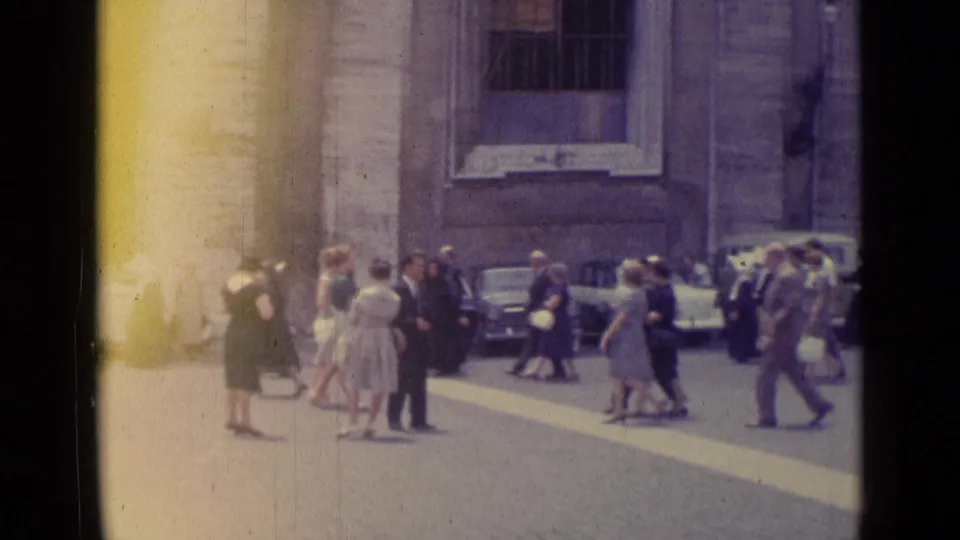 1962 Vatican City frame showing people crossing a bright paved square near parked cars and a stone building facade.