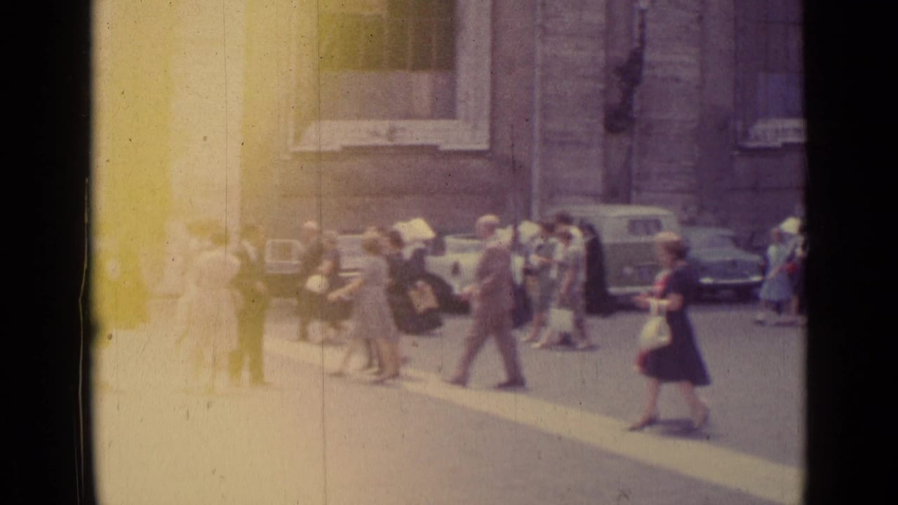 1962 Vatican City home-movie frame showing pedestrians near stone buildings and parked cars, with heavy flare at the left edge.