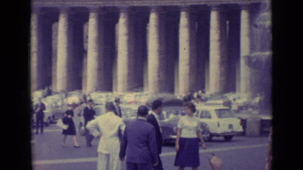 1962 Vatican City frame with pedestrians crossing before the colonnade, surrounded by parked cars and visitors in period dress.