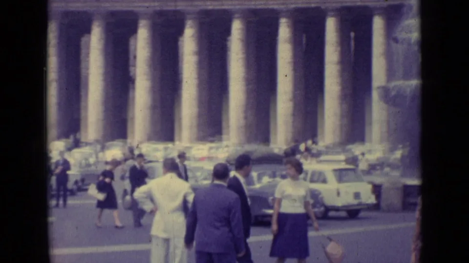 1962 Vatican City frame with pedestrians crossing before the colonnade, surrounded by parked cars and visitors in period dress.