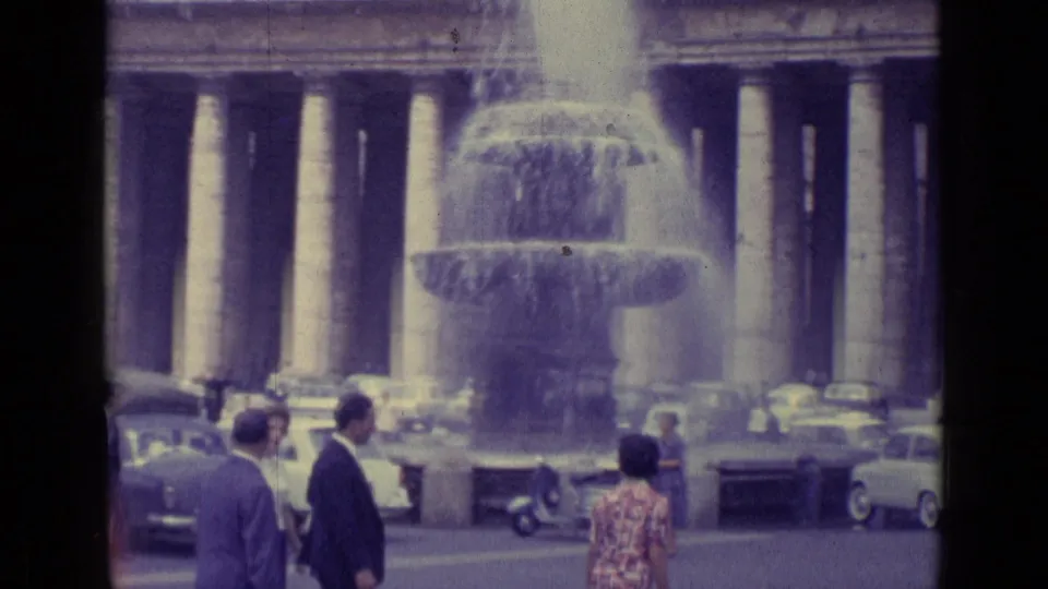 1962 Vatican City 8mm frame showing a large fountain, visitors, cars, and the surrounding colonnade.