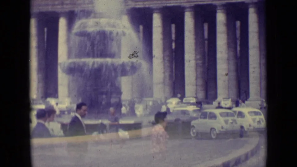 1962 Vatican City street scene with pedestrians near a large fountain, classical columns, and vintage cars around a plaza.