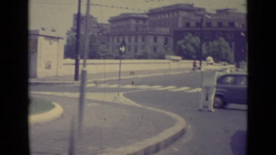 1962 Vatican City roadway view with a traffic policeman in white directing cars near formal buildings and street signs.