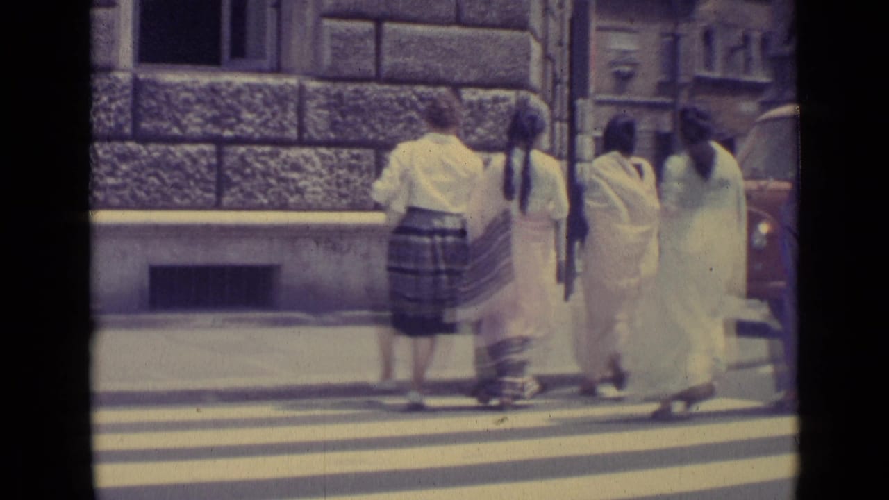 In Vatican City in 1962, women in long traditional South Asian garments cross a zebra crossing beside a stone urban facade.