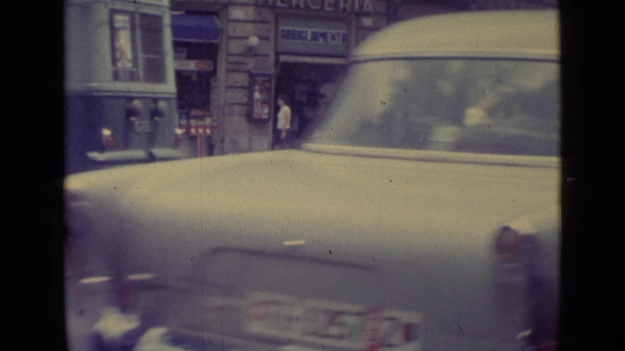 Soft 8mm street view with a pale car passing a shopfront and bus at the edge of frame.
