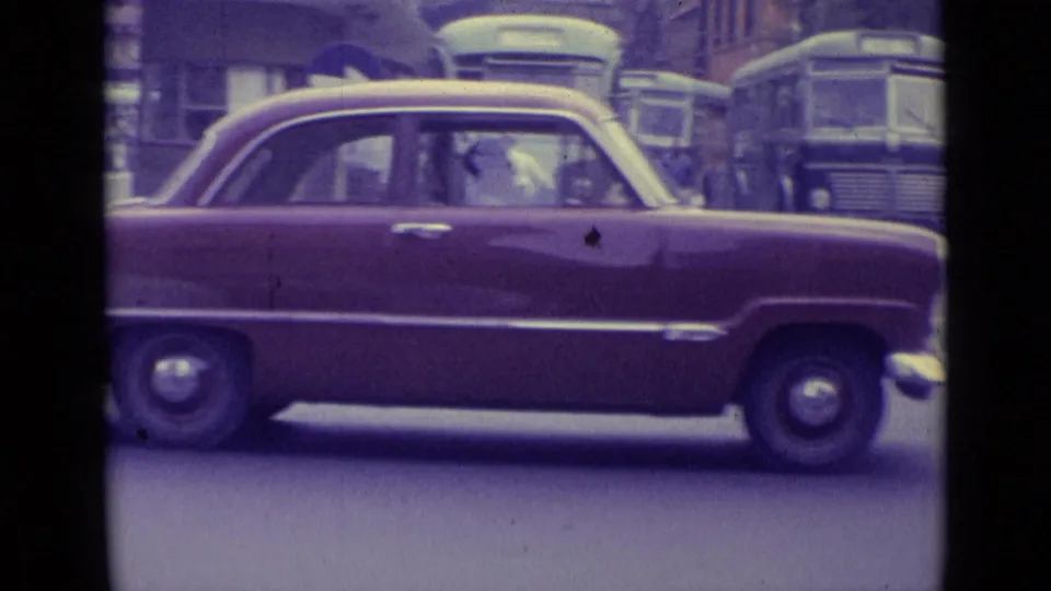 1962 Vatican City street frame with a maroon sedan crossing the foreground and buses visible behind it in heavy traffic.