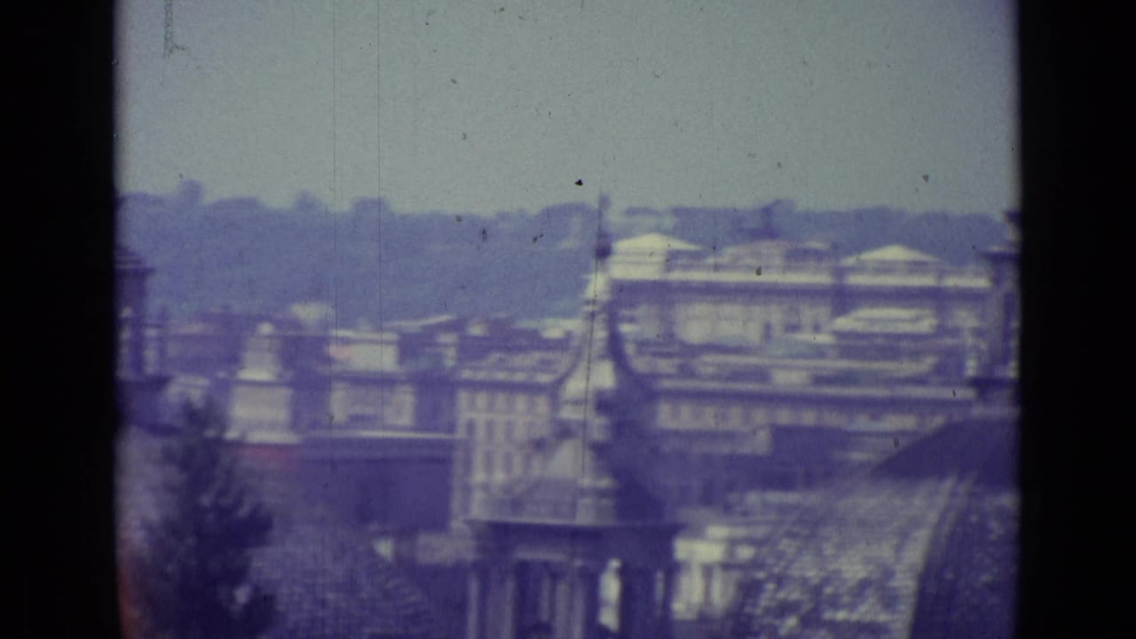 1962 8mm Vatican City rooftop view with hazy buildings, domes, spires, and visible film gate edges.