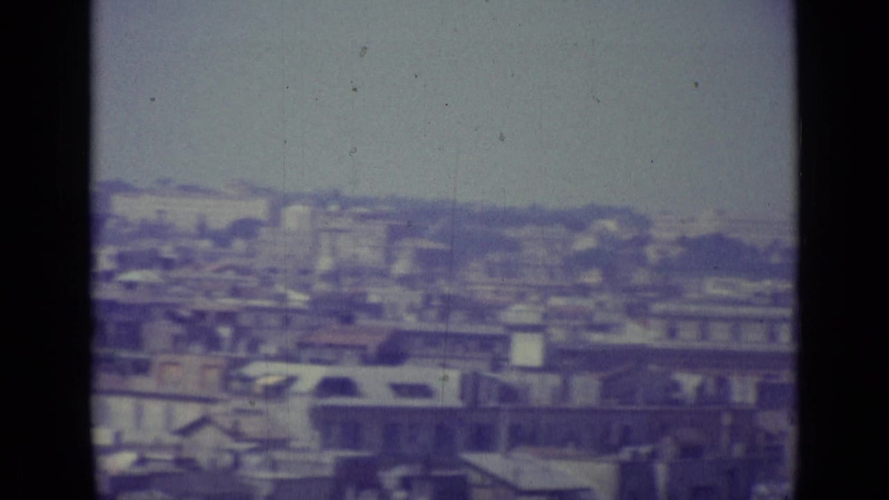 1962 8mm view over Vatican City rooftops, with pale masonry buildings, soft focus, and visible frame division.