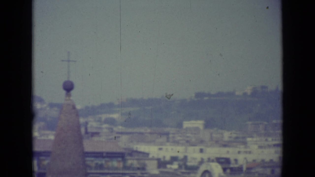 Hazy elevated view with a church cross, distant hillside, buildings, and cars below.