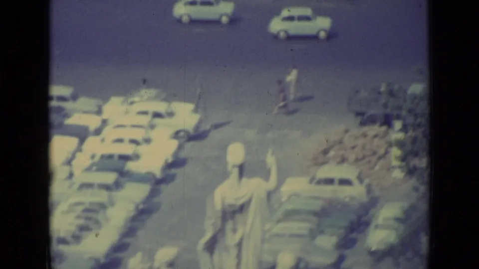 1962 Vatican City home-movie frame with a large statue, rows of parked cars, and small figures near the road.