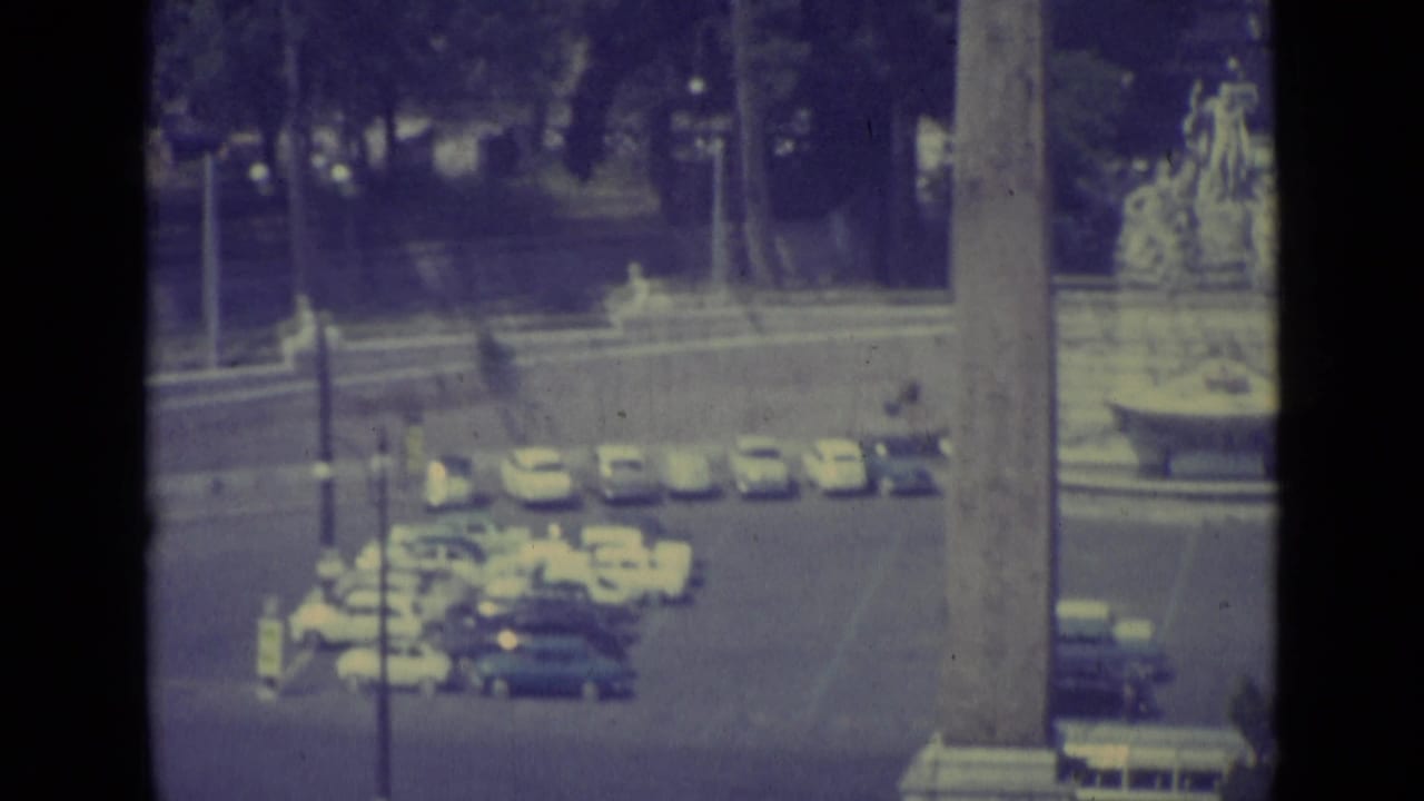 1962 Vatican City home-movie frame showing parked cars, roadway, trees, and a tall vertical monument or structural edge.