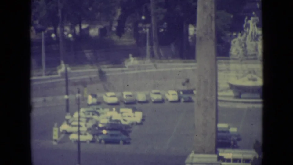 1962 Vatican City home-movie frame showing parked cars, roadway, trees, and a tall vertical monument or structural edge.