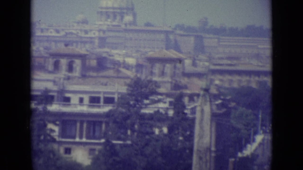 Faded 1962 8mm view of Vatican City with St. Peter's dome, urban rooftops, trees, and a tall obelisk-like monument.