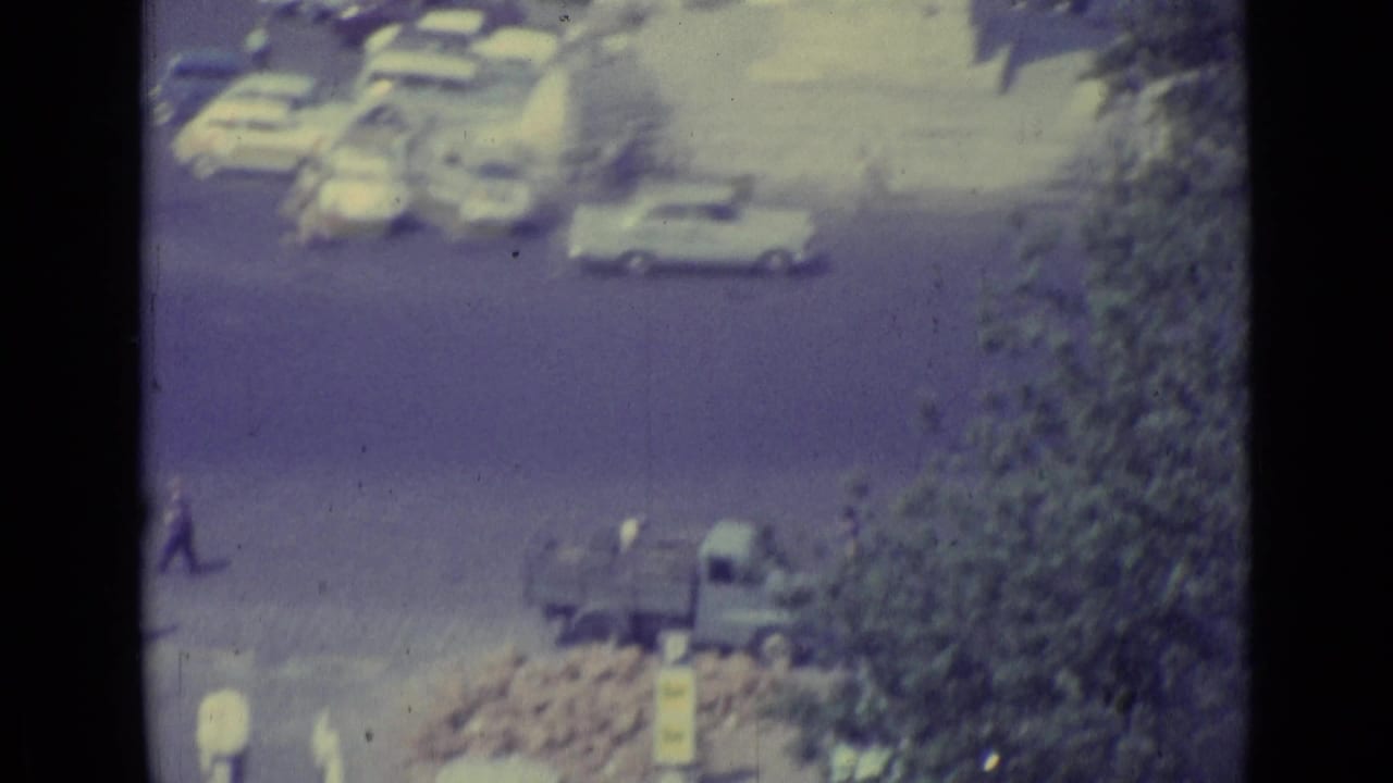 Cars and a small truck move near trees and a paved square, partly obscured by a dark vertical edge.