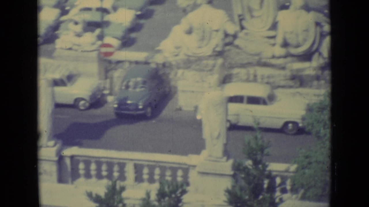 1962 Vatican City home-movie frame showing a sunlit courtyard with classical statues, cars, a red stop sign, and stone railing.