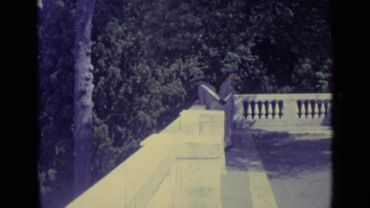1962 Vatican City frame with a suited man in a hat walking along a pale stone balustrade beside a tree-filled garden.