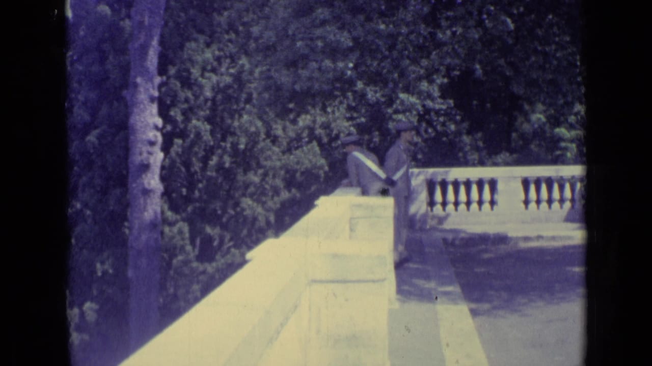 1962 Vatican City home-movie frame showing a stone railing, shaded trees, and suited men moving along a garden-side walkway.