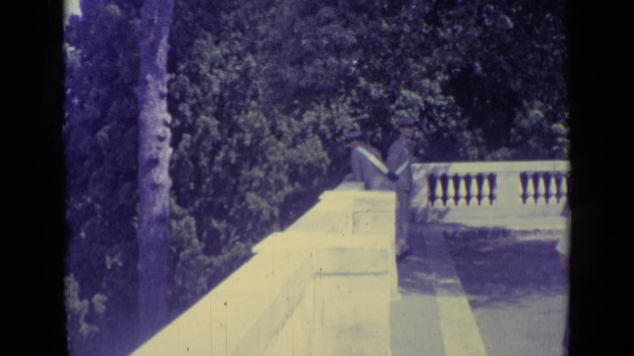 1962 Vatican City frame showing two men in hats and suits near a pale stone balustrade, with dense garden foliage behind.