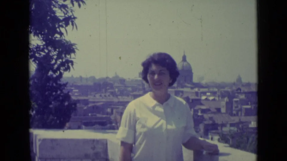 Woman smiles toward the camera on a Vatican City terrace, framed by trees and a distant Roman skyline.
