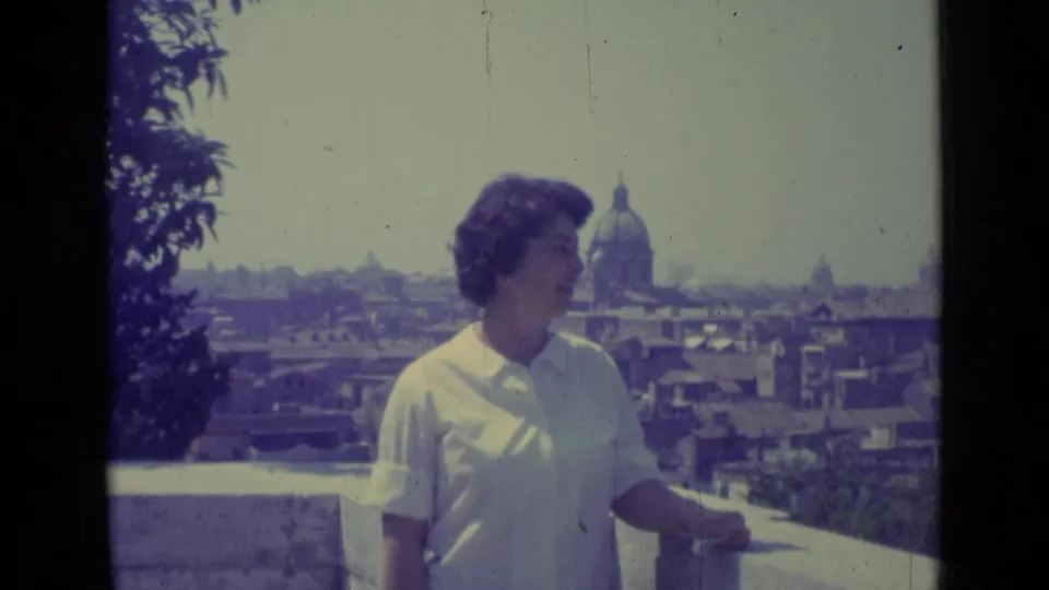 Woman looks across the Vatican City view from a stone terrace, with St. Peter's Basilica dome beyond the rooftops.