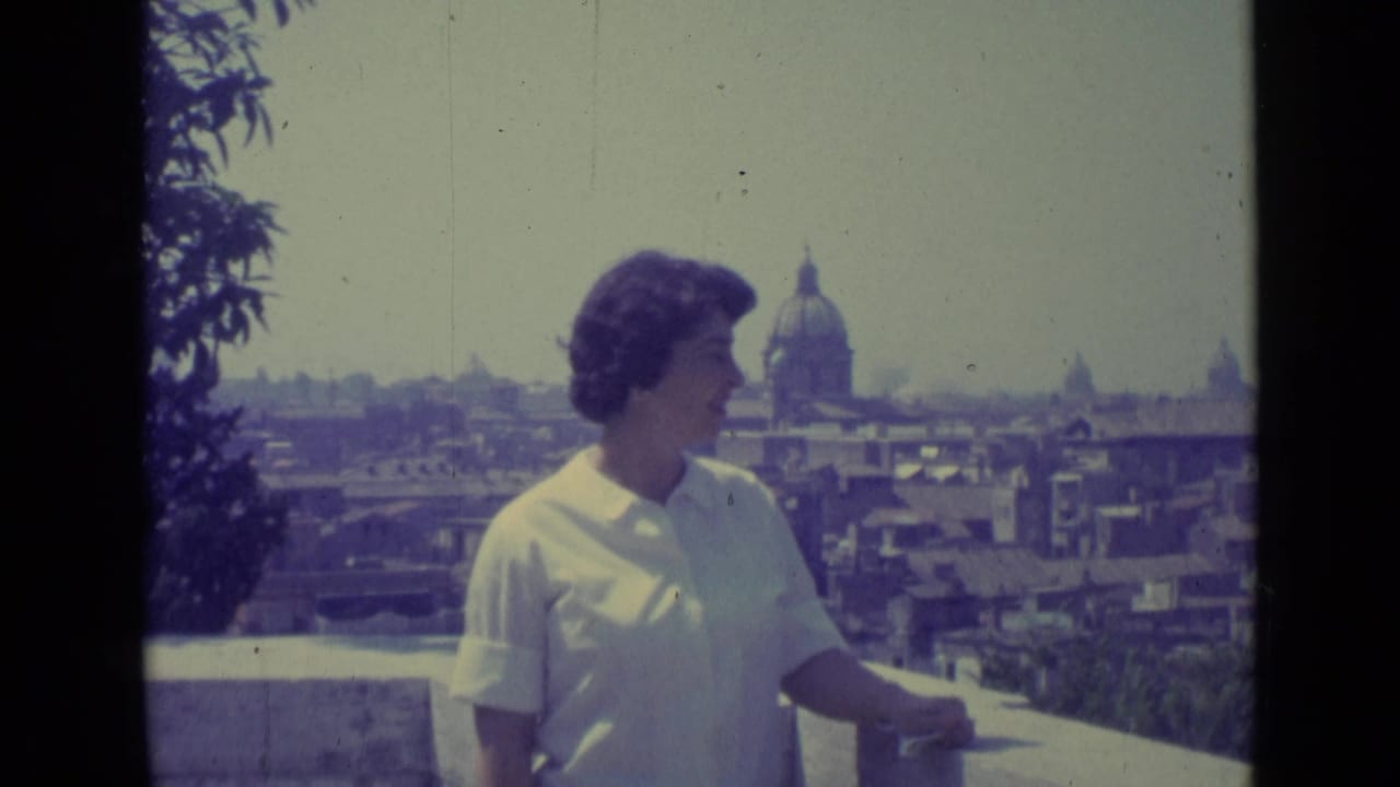 In 1962 Vatican City footage, a woman in a white collared shirt stands on a balcony before rooftops and a large dome.