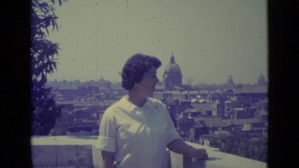 In 1962 Vatican City footage, a woman in a white collared shirt stands on a balcony before rooftops and a large dome.